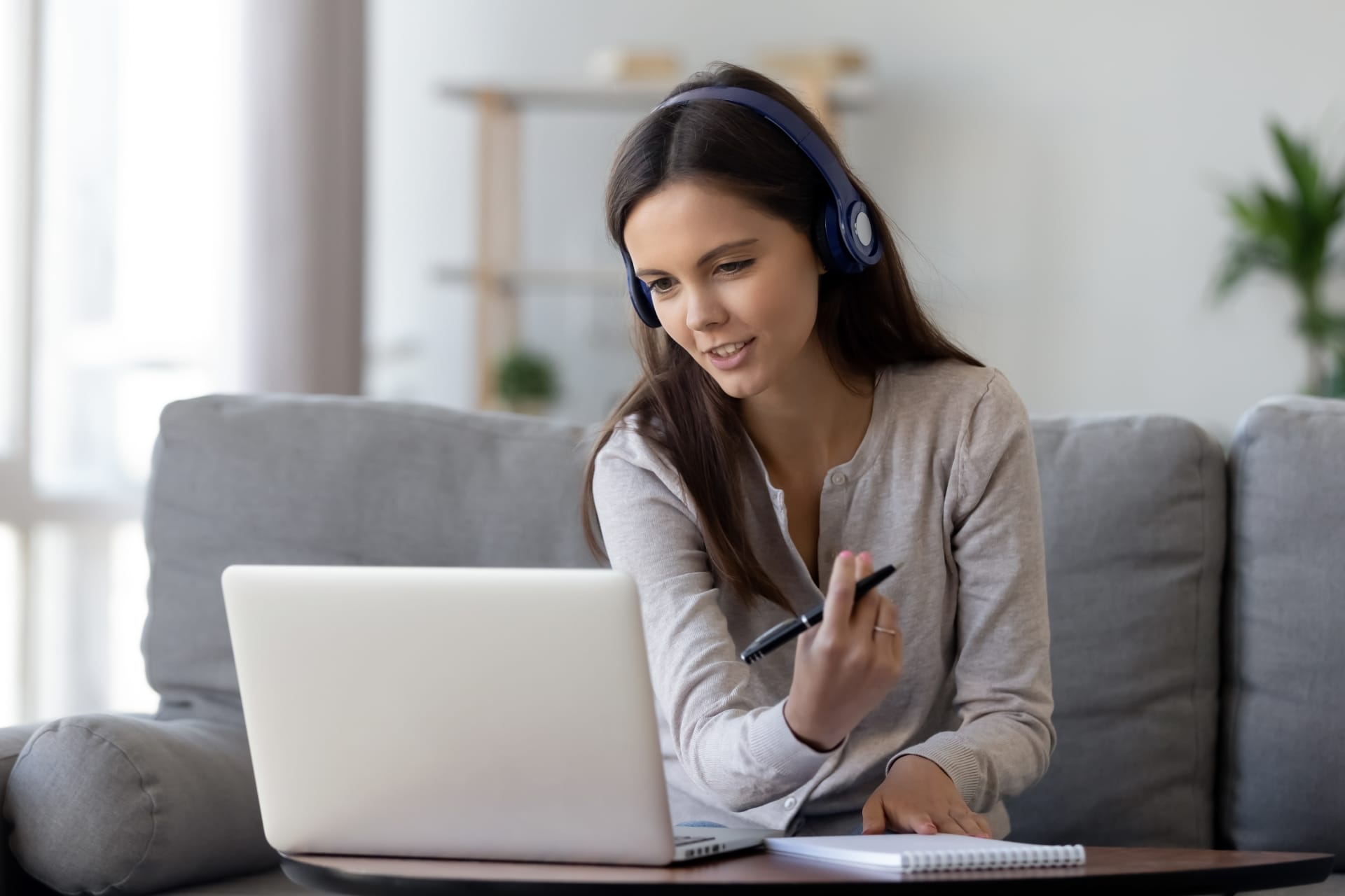 Young woman in headphones doing online course speaking and looking at laptop and making notes